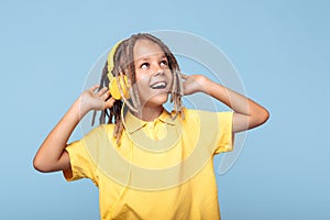 Joyful little boy with african dreads listening music in headphones has pleasant smile over blue background.