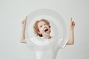 Joyful little boy with curly ginger hair and freckles isolated on white background, pointing upside with happy silly