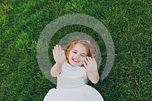 Joyful little cute child baby girl in light dress lie on green grass lawn, showing palms on camera in park. Mother