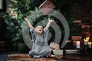 Joyful little girl with blonde curly hair wearing a warm sweater throws up a gift box while sitting on a floor next to