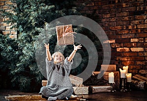 Joyful little girl with blonde curly hair wearing a warm sweater throws up a gift box while sitting on a floor next to