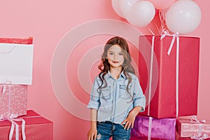 Joyful little girl in blue shirt smiling to camera on big giftboxes with balloons background. Pink colors, birthday mood