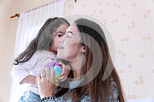 Joyful little girl holding small color ball toy in her hand while kiss her mother in the bedroom. Smile cheerful daughter
