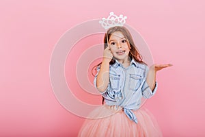 Joyful little girl with long brunette hair in tulle skirt holding princess crown on head isolated on pink background