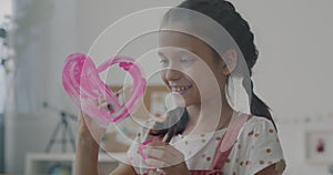 Joyful little girl painting heart on glass board using pink paint and brush smiling indoors at home