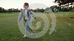 Joyful little girl playing with soap bubbles while standing in green field at sunset, kid spending leisure time outdoors