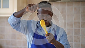 Joyful male African American cook in apron looking at camera imitating fighting with cooking utensils smiling. Cheerful