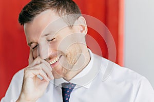 Joyful male in white shirt giggles at funny joke, closes eyes with pleasure. Receives pleasant news. Poses against red wall.