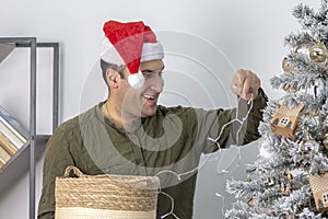 A joyful man of 30-35 years of oriental appearance in a red hat decorates a Christmas tree with a garland and lights.