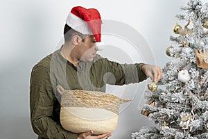 A joyful man of 30-35 years of oriental appearance in a red hat hangs toys from a wooden basket on the Christmas tree.