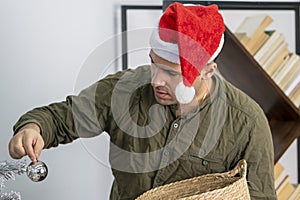 A joyful man of 30-35 years of oriental appearance in a red hat hangs toys from a wooden basket on the Christmas tree.