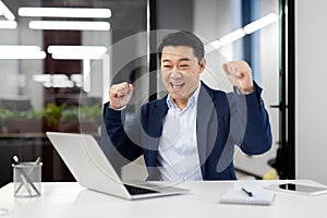 Joyful man dancing at workplace, successful asian businessman in business suit sitting at workplace with laptop inside
