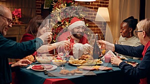 Joyful man disguised as Santa Claus clinking champagne glass with family enjoying Christmas dinner