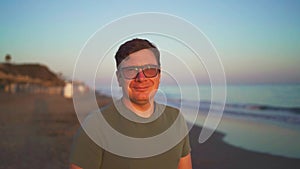 A joyful man with glasses smiling against a blurred beach background during sunset, radiating happiness and relaxation