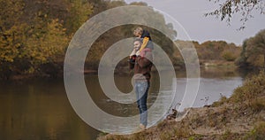 Joyful man is holding child boy on shoulders and enjoying nature of river and forest at autumn, happy father and son at
