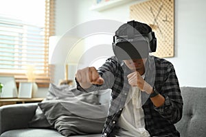 Joyful man wearing virtual reality headset playing simulation boxing game on weekend at home.