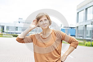 A joyful middle-aged redhead woman in casual clothes stands in the street with her palm to her ear