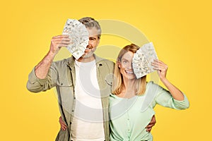 Joyful middle aged spouses hugging and covering eye with bunch of money dollar banknotes, lucky couple holding cash