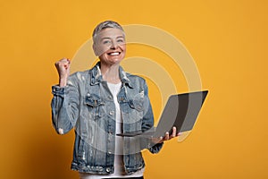 Joyful Middle-Aged Woman Holding Laptop, Celebrating Success Over Yellow Background