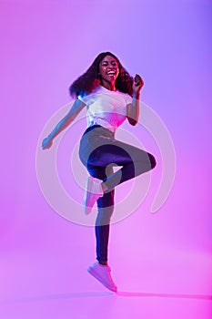 Joyful millennial African American woman flying in air, jumping, smiling at camera in neon light, full length portrait