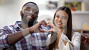 Joyful mixed-race couple showing heart sign made with hands at camera, love