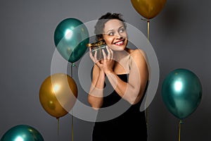 Joyful mixed race pretty woman with beautiful smile holds a gift box near her face, poses on gray background with golden green air