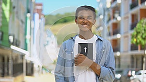 Joyful mixed-race young guy holding holy bible and smiling on cam, religion