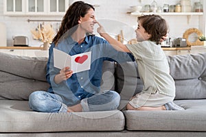 Joyful mom laughing play with cute son hold gift postcard on birthday or mother day sitting on couch