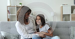Joyful mom tickling excited laughing little girl on home couch