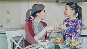 Joyful mother and cute cheerful daughter have fun grimacing silly with vegetables while cooking in the kitchen at home