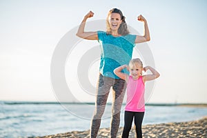 Joyful mother and daughter in fitness gear on beach flexing arms