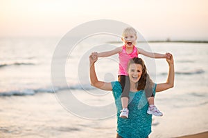 Joyful mother holding daughter on shoulders on beach at sunset