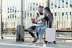 Joyful multiracial male and female mates sitting on tram stop with their suitcases and making photos of their passports.