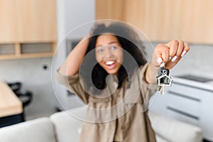 Joyful multiracial woman holding keys with trinket in shape of little cute house and showing it to the camera, selective