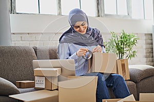 Joyful Muslim woman packaging items into boxes and putting adhesive tape