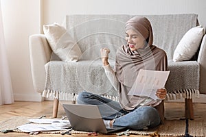 Joyful Muslim Woman Working With Papers And Laptop At Home, Celebrating Success