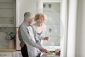 Joyful older retired couple in aprons cooking together in kitchen