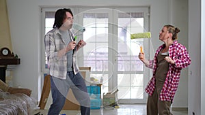 Joyful plus-size Caucasian young couple dancing singing in paint roller brushes in slow motion. Cheerful happy man and