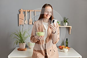 Joyful positive woman with brown hair wearing beige jacket standing in home kitchen interior having breakfast and using mobile