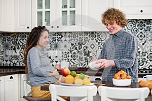 Joyful pregnant woman with dreadlocks drinks tea and watching her curly husband cooking breakfast for her in kitchen