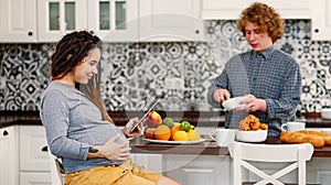 Joyful pregnant woman with dreadlocks drinks tea and watching her curly husband cooking breakfast for her in kitchen