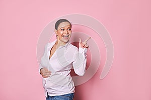 Joyful pregnant woman stroking her belly, smiling looking away, points her finger at copy ad space on pink background