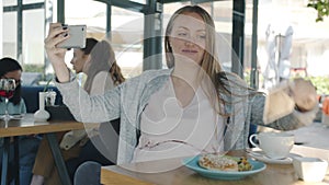Joyful pregnant woman taking selfie having fun in modern summer cafe sitting at table alone