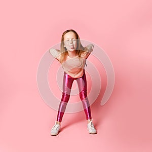Joyful preschool girl in a stylish blouse and leggings is teasing someone by leaning forward, grimacing and wallowing. Full length