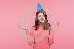 Joyful pretty brunette woman in funny party cap smiling and spreading hands saying surprise, festive mood, party time