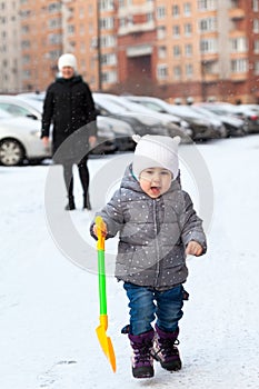 Joyful pretty toddler child running from mother with shovel toy, winter season, people walk on urban parking lot