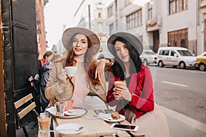 Joyful red-haired girl drinking tea while talking with friend. Brunette young woman spending time in restaurant with her