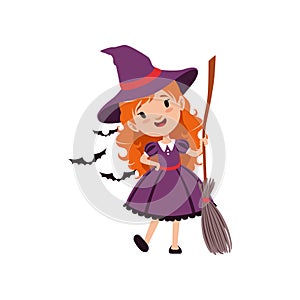 Joyful red-haired girl witch standing with broom and wearing purple dress and hat. Kid character in costume surrounded