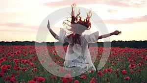 Joyful red-haired woman jumps up in flowered poppy field at sunset, slow motion