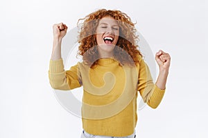 Joyful, redhead curly woman celebrating win, feeling lucky and relieved, yelling yes, achieve success, victory